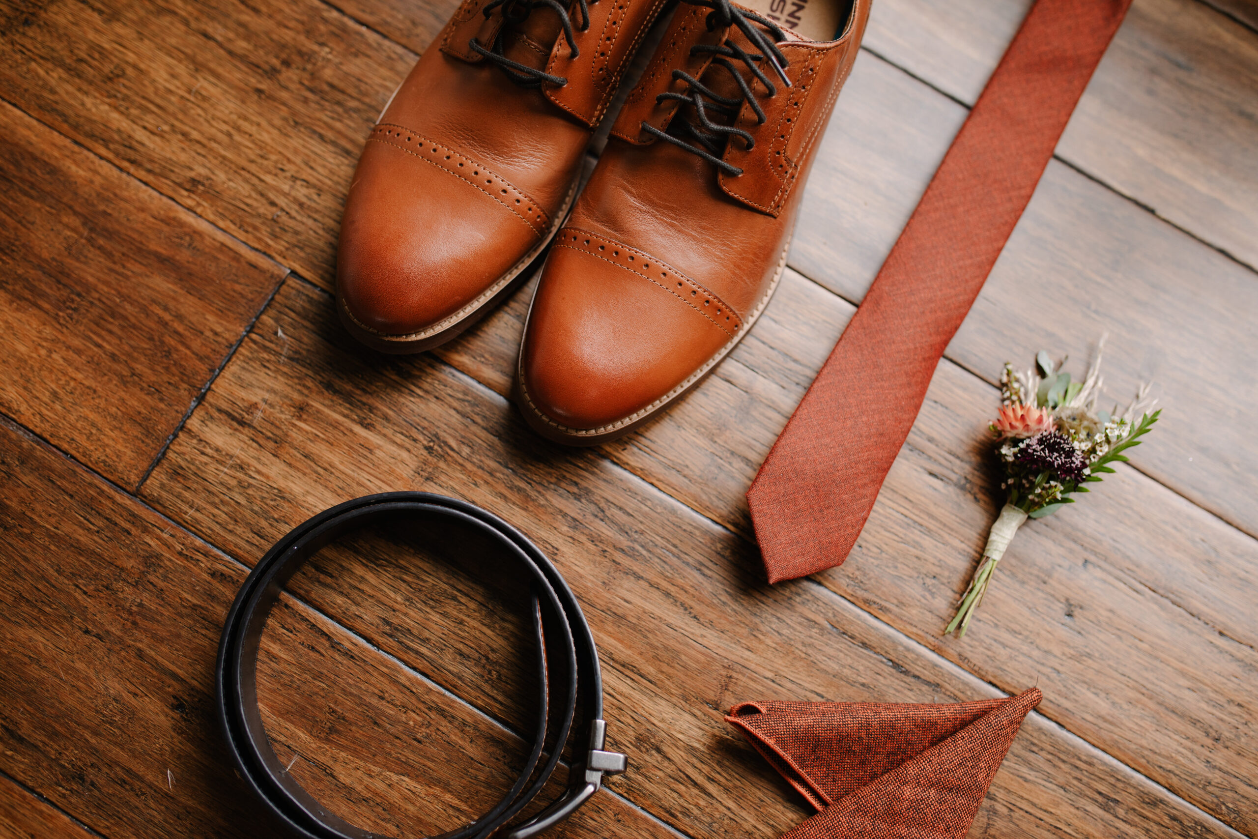 flat lay of groom details including his shoes, tie, belt, flowers and pocket square on a wood floor