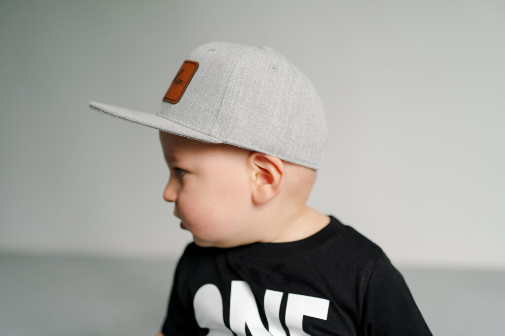 toddler in grey hat looking to the side with a black shirt with "one" written on it in front of a white wall and grey floor